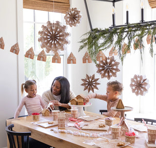 A woman and two children decorate gingerbread houses at a table. The room is adorned with festive paper decorations, greenery, and My Mind’s Eye Gingerbread Reusable Straws featuring a cute gingerbread man pattern.