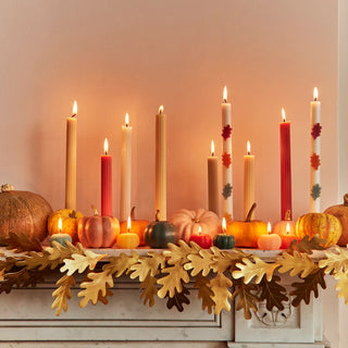 Lit candles of different heights and colors are beautifully arranged alongside small pumpkins and the Gold Oak Leaves Garland by Meri Meri, creating a charming Thanksgiving display on the mantel.