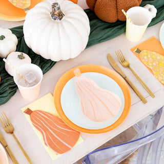 A place setting with a pear-shaped paper design on a die-cut plate, gold utensils, Jollity & Co's Gourd Large Napkins, and a drink with a striped straw, surrounded by pumpkins and fall-themed decor.