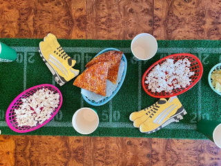 A table with food items, including two pizza slices, popcorns, and chips in baskets, is placed on a football field-themed tablecloth with paper cups around. The Grass Football Table Runner by Talking Tables adds an extra touch to this perfect football party decoration.