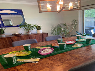 A dining table set for a Super Bowl party features snacks in green and yellow baskets, utensils, napkins, and cups labeled "CHEERS". A Grass Football Table Runner by Talking Tables and plants in the background complete the festive football party decor.