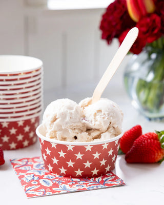 A bowl with star patterns containing two scoops of vanilla ice cream, served with a wooden spoon, on a HAMPTONS FLORAL PAPER COCKTAIL NAPKIN from My Mind's Eye, with strawberries nearby.