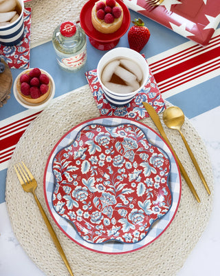 A festive table setting featuring a red and white patterned plate on a woven placemat, with gold cutlery, a cup of tea, and various desserts arranged around it, complemented by My Mind's Eye Hamptons Floral Paper Cocktail Napkins.