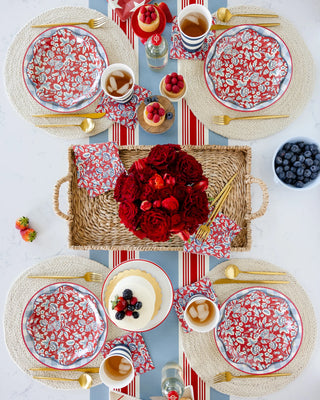 Overhead view of a dining table set with four My Mind's Eye Hamptons Chambray Gingham paper plates, matching cups, golden cutlery, fruits, and a centerpiece with red roses on a wicker tray.