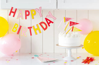 A birthday scene with a "Happy Birthday" banner, a white frosted cake on a stand with decorative flags, festively adorned gold foil accents, My Mind’s Eye Pink Birthday Hat Shaped Plates, pink and yellow balloons, and scattered confetti on a white table.