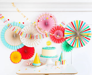 Colorful birthday party decorations with HIP HIP HOORAY PARTY FANS, confetti, and a cake on a table. (Brand Name: My Mind’s Eye)