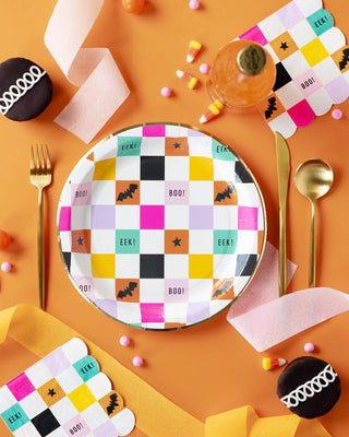 A decorated Halloween-themed table setting with a colorful patterned plate, golden cutlery, cupcakes, candy corn, My Mind’s Eye Halloween Checkerboard Scallop Cocktail Napkin, and a bottle of drink on an orange background.