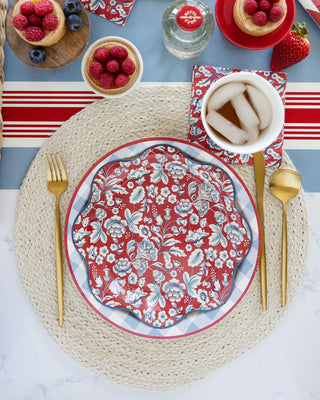 A patterned red and white plate on a woven placemat with gold utensils creates an inviting Americana table setting. Nearby, bowls of raspberries and a cup of iced tea are perfectly placed atop the Hamptons Striped Paper Table Runner by My Mind’s Eye.