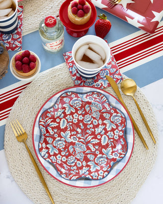 A dining table setting includes a floral red and white plate, gold utensils, a drink, and raspberry-topped desserts. A straw mat and the Hamptons Striped Paper Table Runner by My Mind’s Eye complete this charming arrangement.