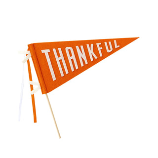An orange triangular flag with the word "THANKFUL" written in white, perfectly serving as a Thanksgiving centerpiece. The Harvest Thankful Felt Pennant Banner by My Mind’s Eye is attached to a wooden pole with ribbon ties, making it ideal for festive table decor.