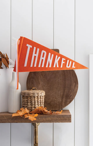 A Harvest Thankful Felt Pennant Banner from My Mind’s Eye is displayed on a shelf with fall-themed decor, including a white vase, a wicker basket, dried leaves, and wooden items. This setup makes for festive table decor perfect for the Thanksgiving season.