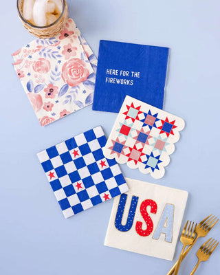 A patriotic table setting featuring a "Here For Fireworks" paper cocktail napkin by My Mind's Eye, quilt-patterned and checkered coasters, a floral napkin, gold cutlery, and a jar with straws.