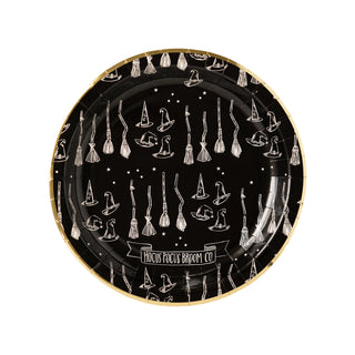 The My Mind’s Eye Hocus Pocus Broom Plate is a round black plate with a gold rim, adorned with white drawings of witch hats, broomsticks, stars, and the text "Hocus Pocus Broom Co." It's perfect for spooky party Halloween decorations.