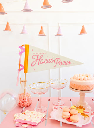 Festive party setup with a "Hocus Pocus" felt banner by My Mind’s Eye, champagne glasses, a cake, macarons, a jar of candy, and party hats hanging in the background—perfect for a magical gathering.