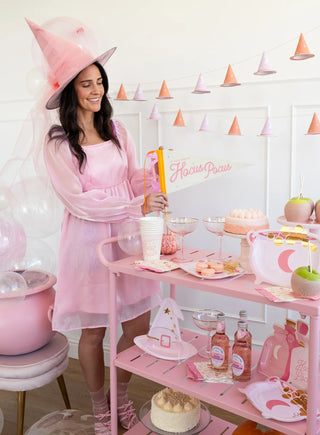 A woman dressed in a pink witch costume stands by a decorated table filled with various pink-themed treats and party items, beneath a garland of pink witch hats. A "Hocus Pocus Felt Banner" by My Mind’s Eye is visible, adding charm to the magical gathering.