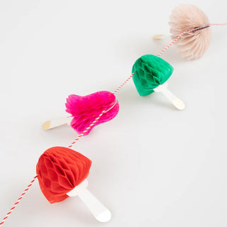 The Honeycomb Mushrooms Mini Garland by Meri Meri, featuring charming paper mushrooms attached to popsicle sticks and strung together on red and white string, is laid out on a white background creating a festive decoration.