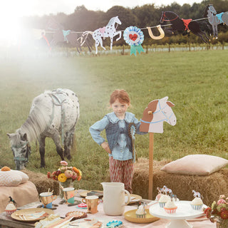 A child stands at an outdoor horse-themed party. A table is set with horse decor and party items, including a Meri Meri Horse Cupcake Kit with charming horse toppers, while a pony grazes in the background.