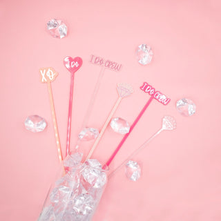 Five decorative "I Do Crew Drink Stirrers" by NPW, bearing phrases like "I DO CREW" and "XO," protrude from a glass filled with ice against a pink background scattered with plastic diamonds—perfect for your bachelorette party.