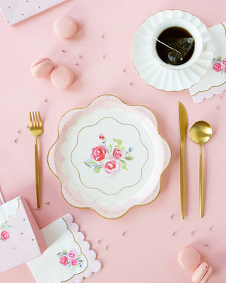 A decorative table setting with a floral-patterned plate, gold-colored cutlery, and a cup of tea, surrounded by pink macarons on a pink background. The scene is elevated with My Mind’s Eye Tea Party Scalloped Round Paper Plate that harmonizes beautifully with the delicate arrangement.