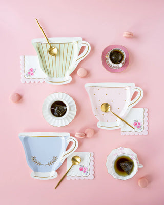 Three decorative tea cups with saucers, gold spoons, and tea bags on a pink surface, accompanied by colorful macarons and **My Mind's Eye Tea Party Rose Corner Paper Cocktail Napkins**, setting the perfect scene for an elegant tea party.