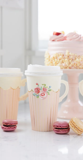 Two floral-patterned Tea Party Handled Paper Cups by My Mind’s Eye with sleeves and lids sit beside a cake stand holding a decorated cake and three macarons on a white counter, perfect for an elegant tea party setup.