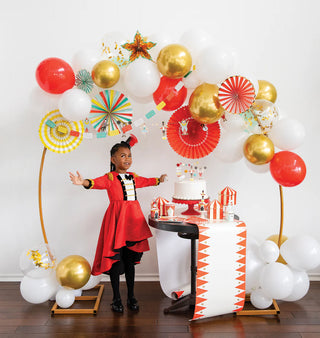 A child in a red circus-themed outfit poses with outstretched arms in front of a decorated table adorned with a whimsical tablescape, including a cake, balloons, paper fans, and a Carnival Paper Table Runner by My Mind’s Eye.