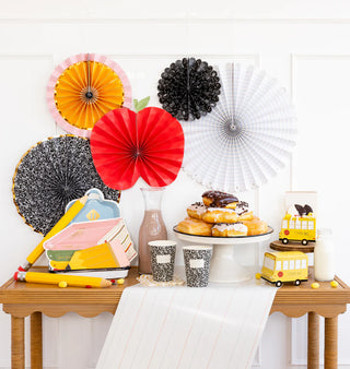 A table is set with pastries, milk, and books. Decorative pinwheels, a school bus toy, a crayon box, notebook-themed cups and paper, and a playful Backpack Plate Set by My Mind’s Eye complete the school-themed decor.