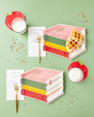 Four book-shaped plates with waffles and gold forks, two apple-shaped drinking cups accompanied by Apple Shaped Napkins from My Mind’s Eye, library cards, and multicolored sprinkles on a green surface. Perfect for back-to-school celebrations!