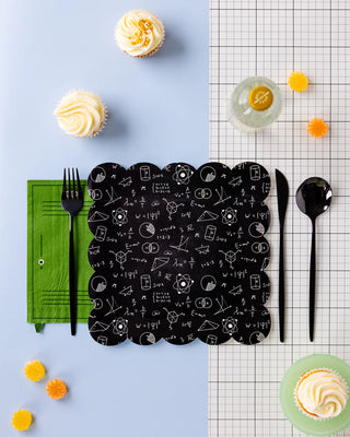 Table setting with a black science-themed placemat, Locker Shaped Napkin by My Mind’s Eye in green with black utensils, bottle with lemon slice, two frosted cupcakes, and candies on a light blue and graph paper-patterned surface—perfect for a school-themed celebration.