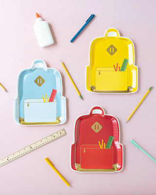 Three Backpack Plate Sets by My Mind’s Eye in yellow, red, and blue are shown with school supplies scattered around, including pencils, a ruler, a glue bottle, and colored pens on a pink surface—perfect for organizing or serving fun school lunches.