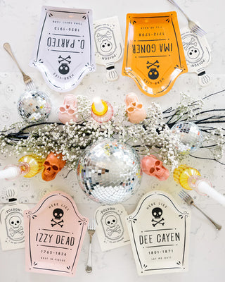 A Halloween-themed table setting featuring plates styled as mock gravestones with inscriptions like “D. PARTED,” “IMA GONER,” “IZZY DEAD,” and “DEE CAYEN.” The decor is enriched with skull decorations, baby’s breath flowers, and disco balls. To add an extra spooky touch, the setting is complemented by Poison Bottle Shaped Paper Dinner Napkins from My Mind’s Eye.