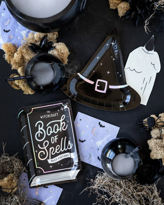 A table set with a Witching Hour Book of Spells Shaped Plate by My Mind’s Eye, a "Book of Spells" notebook, cauldrons emitting smoke, bat-themed napkins with holographic foil accents, and dried moss decorations.