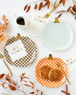 Three plates from the My Mind’s Eye Harvest Gingham Pumpkin Shaped Paper Plate Set with "Give Thanks" cards placed on them, a small pitcher, and autumn leaves arranged on a white surface. The bottom plate, inspired by the gingham-shaped pumpkin designs of the set, holds two pumpkin-shaped pastries perfect for a festive harvest gathering.