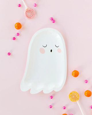 The Trick or Treat Ghost Shaped Plate by My Mind’s Eye, featuring a peaceful expression, is surrounded by colorful lollipops, orange candy balls, and pink candy beads on a light pink background, making it the perfect addition to your party table this Halloween.