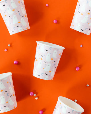 Trick or Treat Party Cups by My Mind’s Eye are arranged on an orange background, surrounded by small pink, silver, and purple beads and star-shaped decorations. These party cups create a festive atmosphere for any spooky gathering with their whimsical ghost prints.