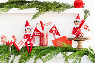 Three elf dolls in red outfits hold festive flags on a shelf adorned with green garland, candy canes, and a jar of red candies, creating the perfect setting for a Christmas party. Consider adding Elf Collar Party Cups by My Mind’s Eye to complete this holiday punch of festive cheer.