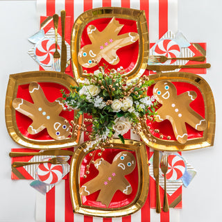 A festive table setting with gingerbread man plates, a floral centerpiece, gold cutlery, and a striped red and white tablecloth showcases Jollity & Co's Red Peppermint Napkins accented with silver foil details.