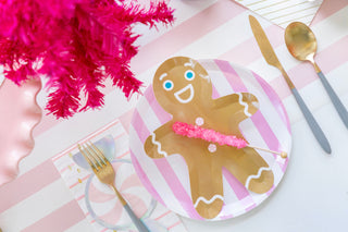 A Jollity & Co Gingerbread Man Plate designed for Christmas parties sits elegantly on a pastel table. Topped with a pink rock candy stick and flanked by a gold fork, knife, and spoon, it's perfect for serving holiday desserts in style.