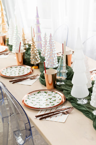 A festive table setting featuring tree-shaped decorations includes Jollity & Co's Holly Print Dinner Plates, complemented by copper cups, striped napkins, and silverware on a light wood table with a green runner.