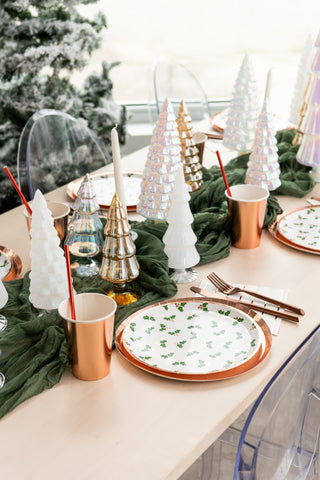 A festive table setting with tree-shaped ornaments, green napkins, and copper accents featuring Jollity & Co's Holly Print Dinner Plates and cups with red straws.