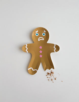 A Gingerbread Man Plate by Jollity & Co, with a frowning face and missing leg, lies on a white surface surrounded by crumbs. It evokes the humorous mishaps that frequently occur at Christmas parties, where these whimsical plates can quickly become part of amusing stories.