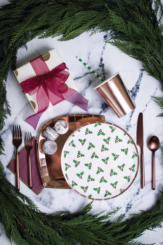 A festive table setting showcasing Jollity & Co's Holly Print Dinner Plates, paired with copper utensils, a cup, and a ribboned gift on a marble surface, all beautifully accented by a lush green garland.