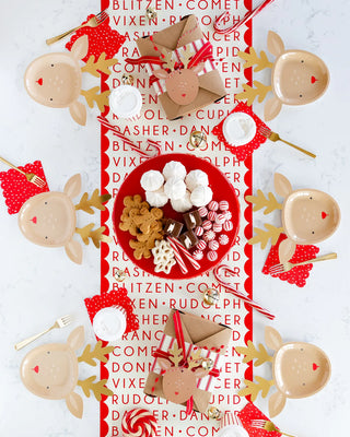 A festive tablescape showcases reindeer plates, candy canes, and milk glasses accompanied by a central plate of cookies and chocolates. The setting is beautifully arranged on the Believe Reindeer Table Runner by My Mind’s Eye in vibrant red and white.