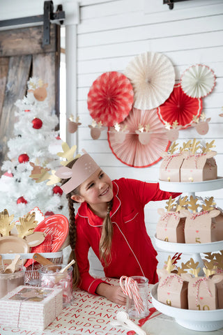 A child in red pajamas and an antler headband stands beside a decorated table with reindeer cupcakes, a white Christmas tree, and a festive tablescape featuring the Believe Reindeer Table Runner by My Mind’s Eye.