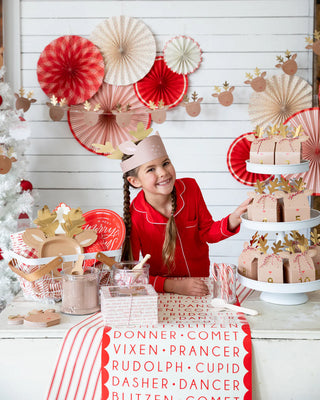 A girl in red pajamas stands at a festive holiday dessert table displaying cakes, with decorations featuring red and pink paper fans, a Believe Reindeer Table Runner by My Mind’s Eye adorned with reindeer names, and playful reindeer cups.