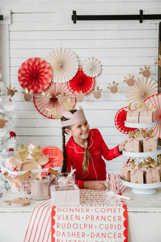 A girl in red pajamas stands by a festive tablescape adorned with reindeer decorations and the charming Believe Reindeer Table Runner by My Mind’s Eye. Brown paper-wrapped gifts sit nearby, with a white Christmas tree glowing in the background.