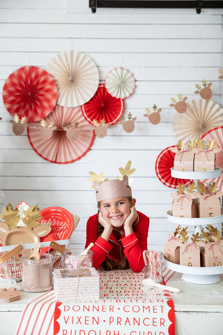 In a festive setting, a child with reindeer antlers joyfully surveys the vibrant holiday decorations and gifts on a table, which feature the charming Believe Reindeer Table Runner by My Mind’s Eye.