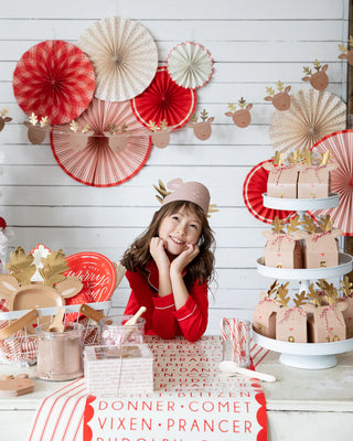 A child, sporting a charming reindeer hat, beams at a festive tablescape decorated for Christmas with the Believe Reindeer Table Runner by My Mind’s Eye, featuring red and white accessories, gift boxes, and pastries.