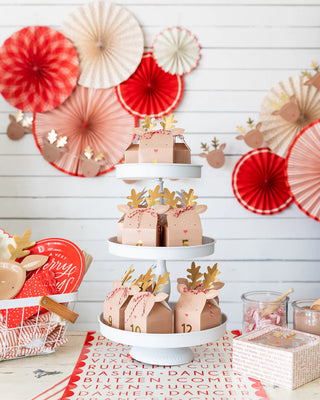 A festive tiered tray showcases reindeer-themed gift boxes, beautifully complemented by the "Believe" Reindeer Table Runner from My Mind’s Eye. Paper fans and reindeer decorations add charm to the background, accentuating the utensils and festive items on a nearby table for a perfect Christmas tablescape.