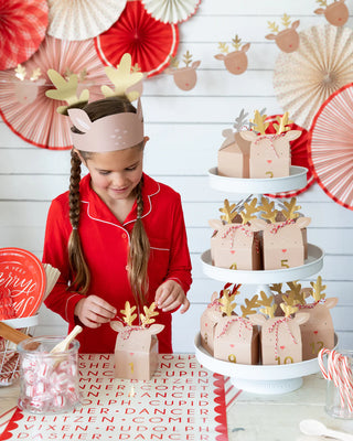 A child in red pajamas decorates the Believe Reindeer Table Runner by My Mind’s Eye with candy canes, creating a festive tablescape surrounded by sparkling decorations.